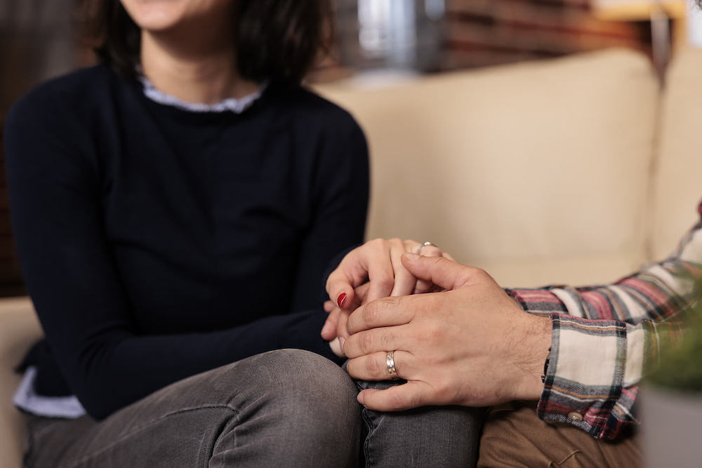 Couple sitting on a couch holding hands while in couples therapy. Discover how couples therapy in Miami, FL can help you overcome your conflict you face in your relationship with the help of a skilled couples therapist.