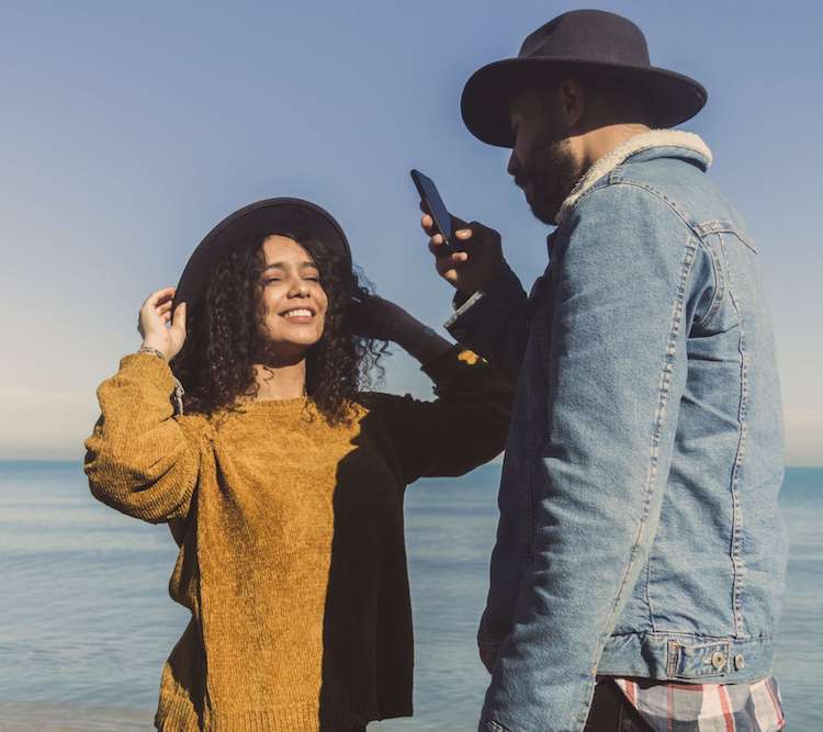 Image of a woman posing near the ocean smiling and wearing a hat while her partner takes the photo on a cell phone. Uncover your communication styles with your partner with the help of a couples therapist in Miami, FL in couples therapy.
