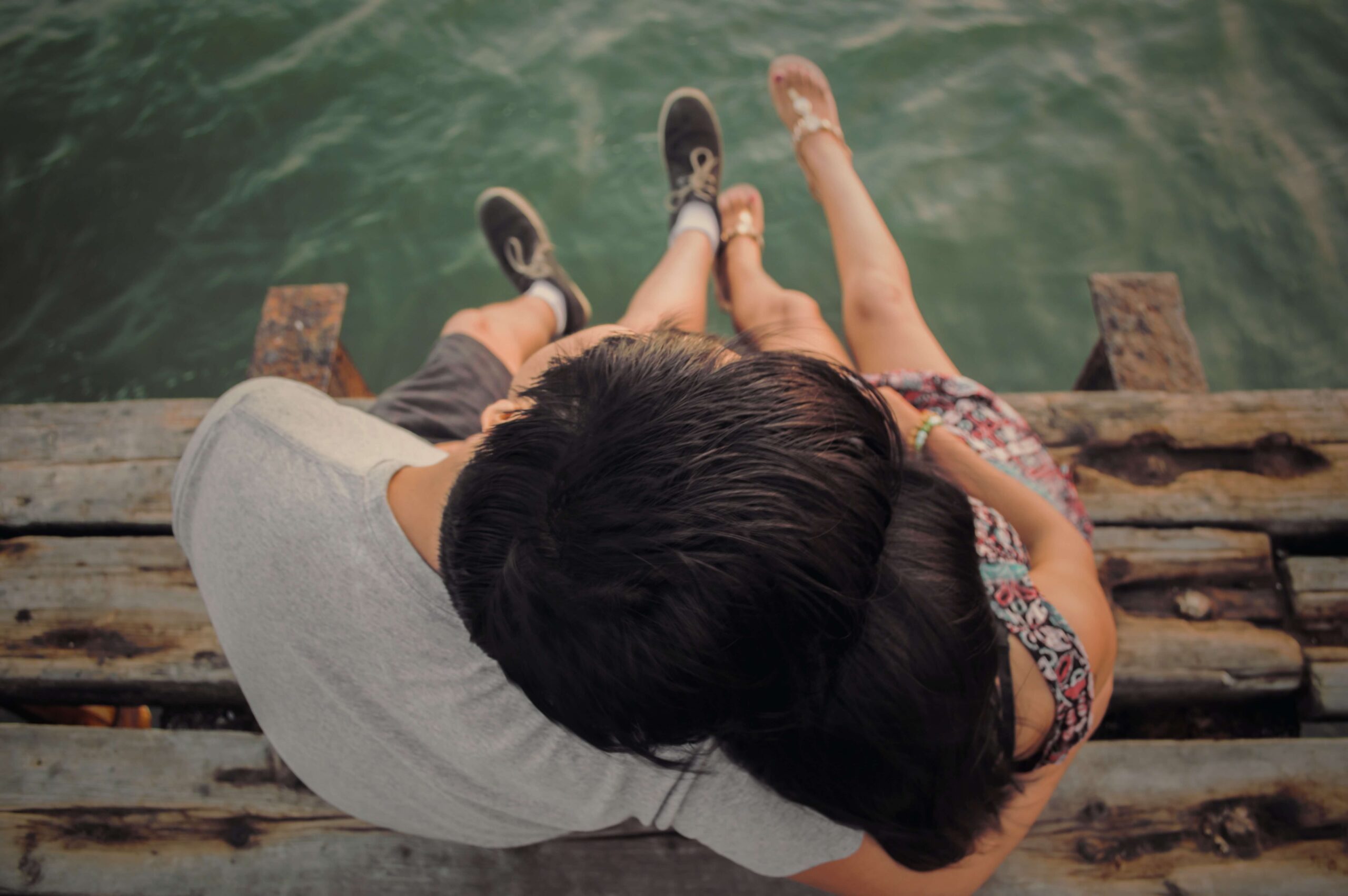 Image of a couple sitting on the side of a pier hugging and dangling their feet over water. With the help of couples therapy in Miami, FL you can overcome your communication issues and improve your connection.