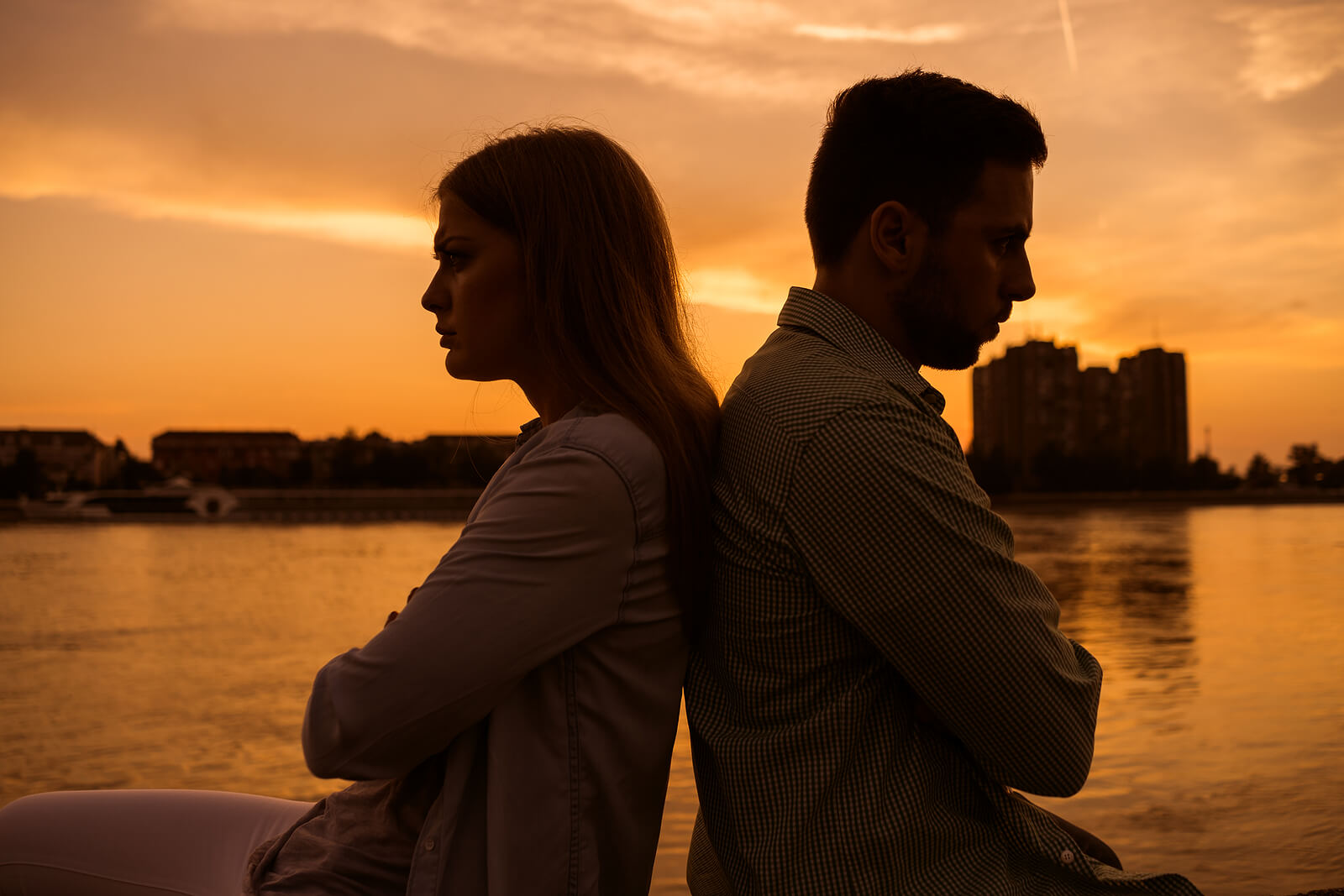 Image of an upset couple sitting back to back next to water while the sun sets. Discover how couples therapy in Miami, FL can help you and your partner overcome your issues and take your mind off of divorce.