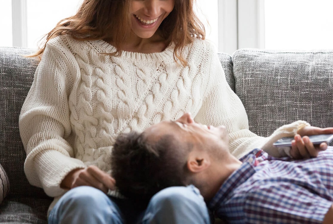 Image of a couple sitting on a couch smiling at each other. Discover how infidelity recovery in Miami, FL can help you and your partner reconnect and adjust with infidelity PTSD.