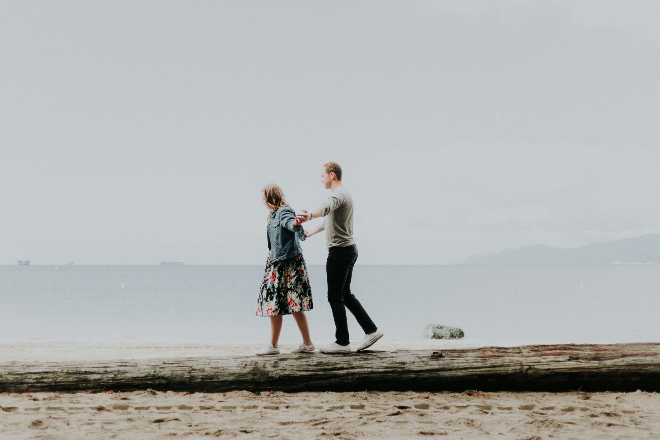 Image of a young woman and man walking on a log on the beach holding hands to balance each other. If you struggle to rebuild trust after cheating in your relationship, learn how couples therapy in Miami, FL can help.