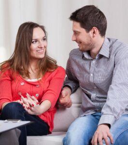 A smiling couple sits close together on a couch, making eye contact and talking openly during a guided session. An example of what makes specialty couples therapy different from what they'd tried before. Out of network couples therapy in Miami Beach, Tampa, and Orlando for couples asking, "should we try couples therapy again?" Book your session today.