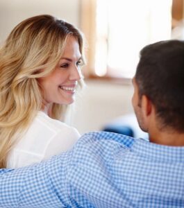 A smiling woman with wavy blonde hair and a white blouse looks warmly at a dark-haired man, whose arm rests around her shoulders in a bright room. EMDR in couples therapy supports infidelity recovery and betrayal trauma healing for couples in Port St. Lucie, Pembroke Pines, and West Palm Beach. Reach out today to start rebuilding your relationship together.