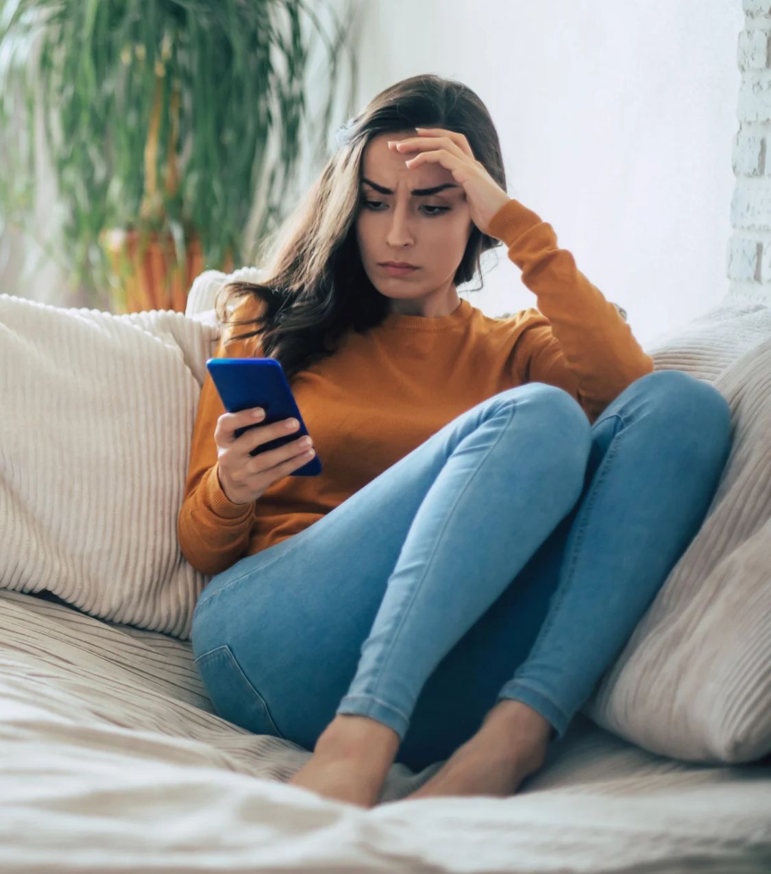 A young woman with long dark hair, sits curled up on a cream sofa with her hand pressed to her forehead as she frowns at her phone. Reliving the affair and affair triggers are common signs of betrayal trauma for hurt partners in Miami, Aventura, and Ft. Lauderdale. Book a free 45-minute consultation with Idit to start healing.