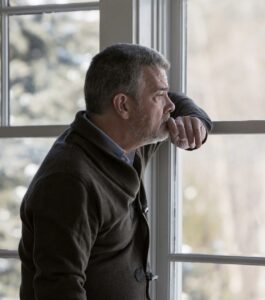 A gray-haired father leans against a window with his fist pressed to his chin, gazing outside with a heavy, distant expression, as if waiting to hear from his grown child. Guidance for when an adult child cuts off a parent after an affair, for staying after an affair, and for healing from parental infidelity in Miami Beach, Tampa, and Orlando. Start rebuilding your family with a Florida infidelity recovery specialist.