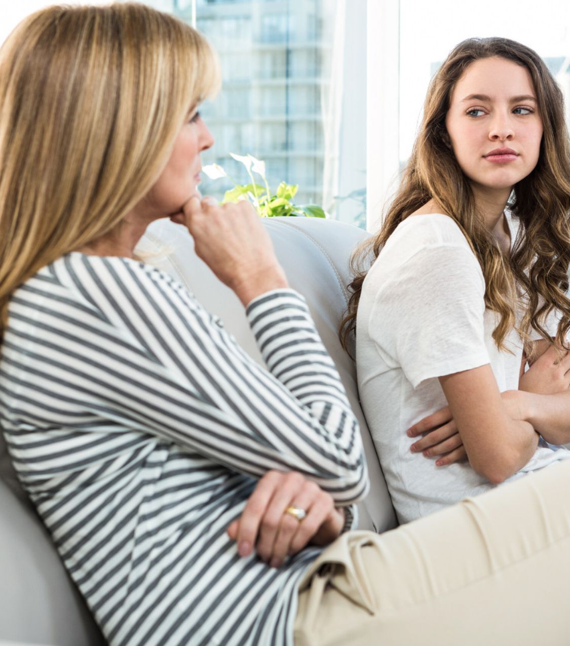 A mother sits on a white sofa with her hand on her chin, looking away, while her young adult daughter sits beside her with arms crossed and a guarded sideways glance. Support for parents navigating adult children after an affair, telling adult children about an affair, and infidelity recovery in Miami, Aventura, and Ft. Lauderdale. Request a private consultation with Idit Sharoni, LMFT.